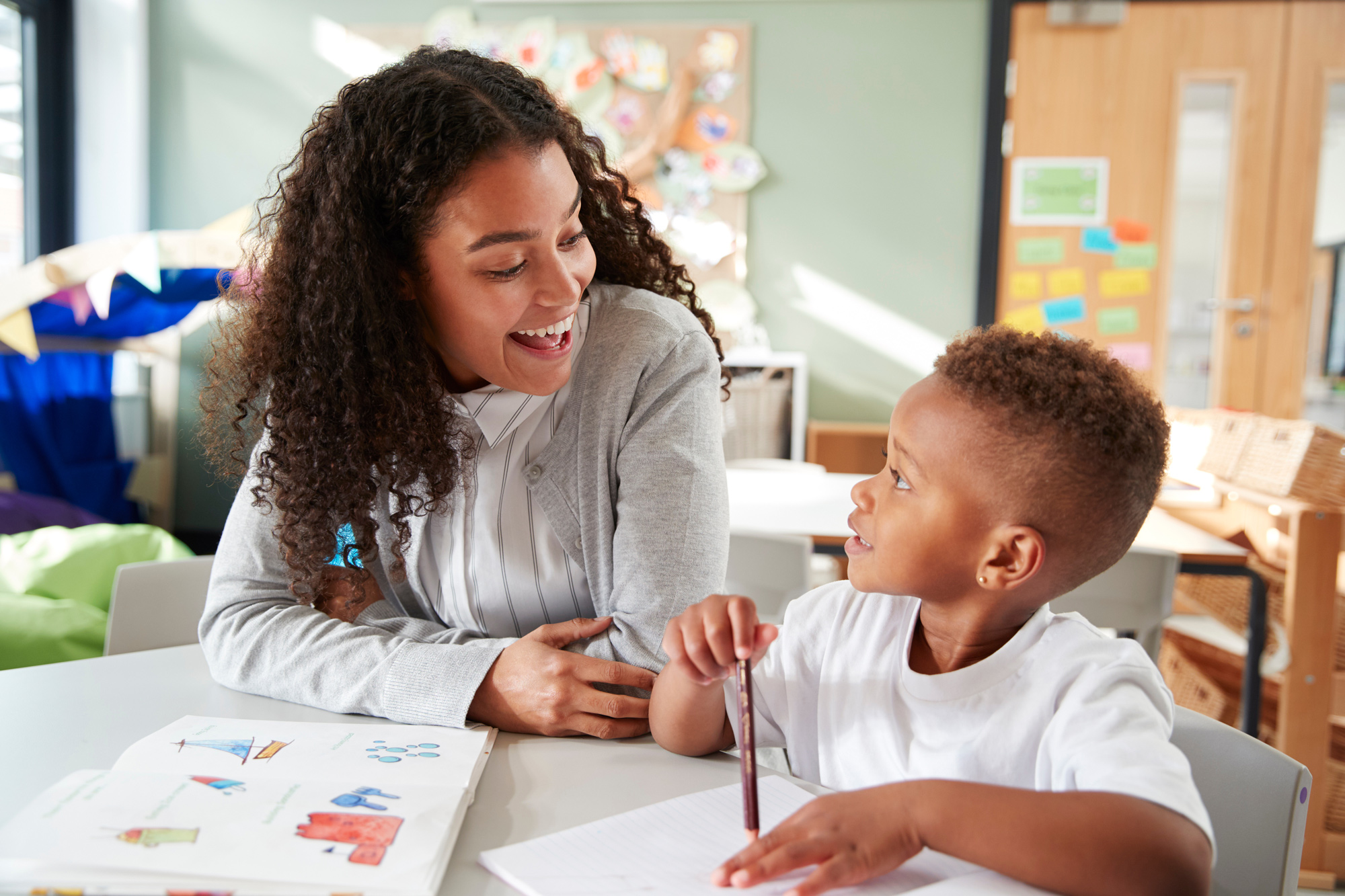 Female infant school teacher working one on one with a young schoolboy, sitting at a table smiling at each other